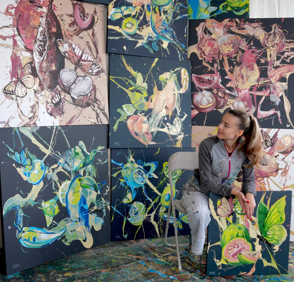 Silvi The Artist seated in her studio surrounded by vibrant abstract paintings inspired by nature, fruit, and movement.
