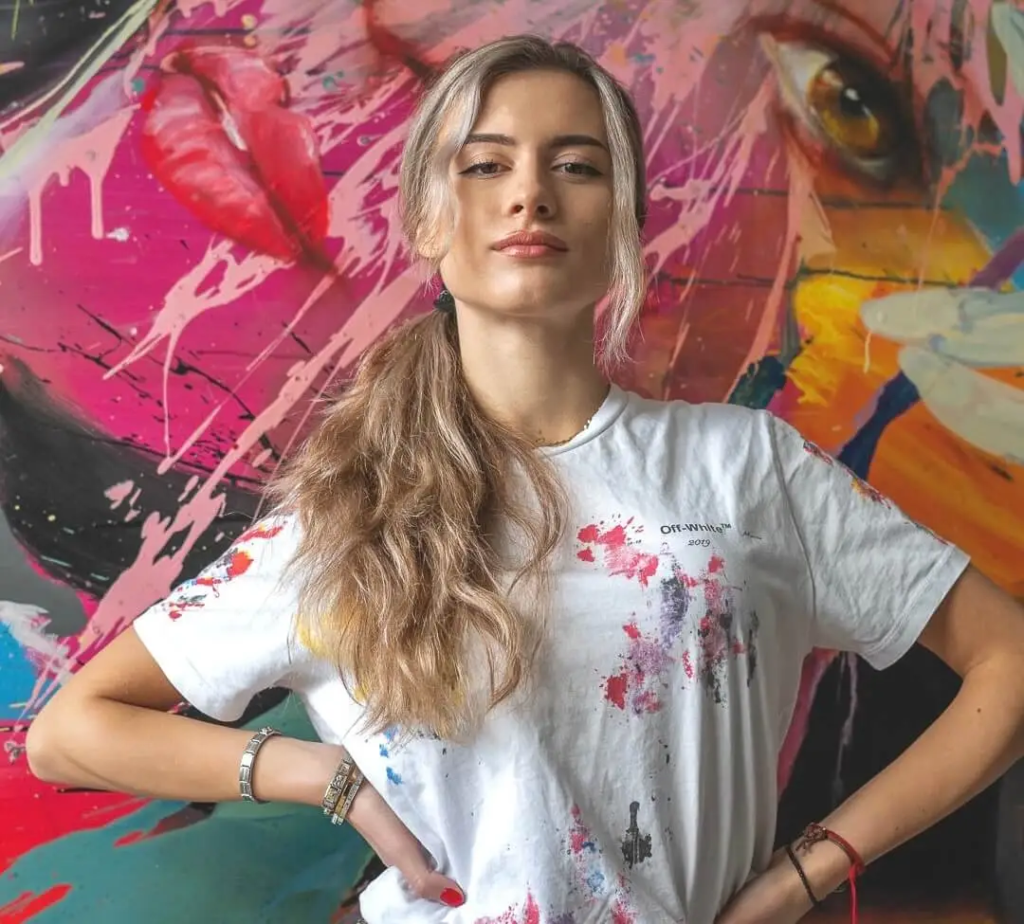 Confident portrait of SILVI The Artist standing in front of her vibrant abstract painting with colorful paint splatters, wearing an Off-White t-shirt covered in artistic drips, long blonde ponytail, hands on hips in her creative studio