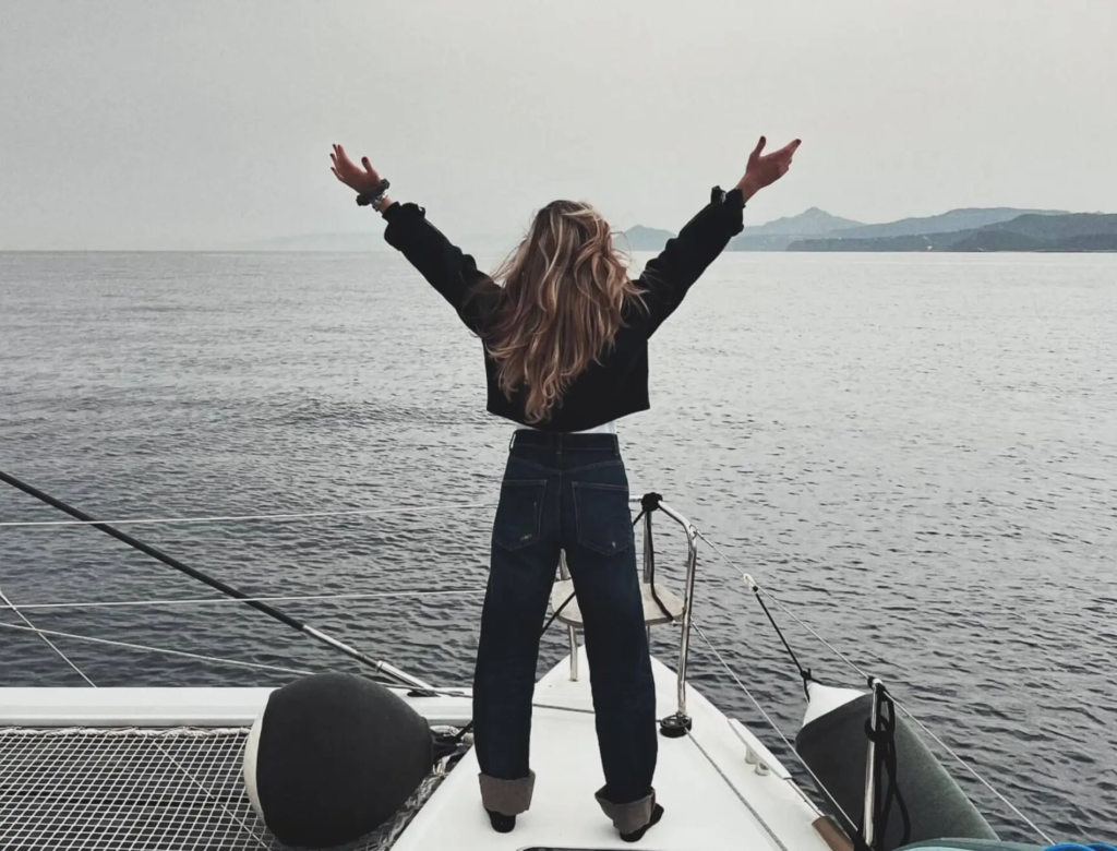 SAILING: Contemporary Mixed Media Painting Series by SILVI (2024/2025) 1 SILVI The Artist standing triumphantly on a sailboat with arms raised wide in freedom, long blonde hair flowing in the wind, overlooking calm open sea and distant misty mountains