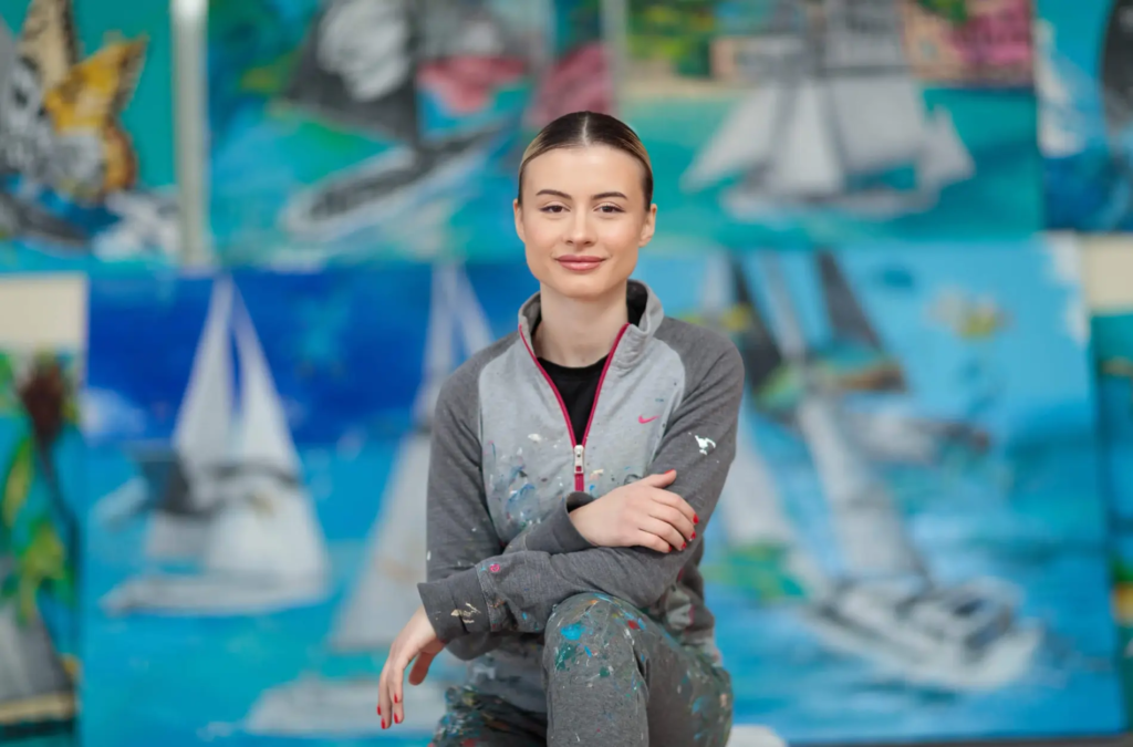 SAILING: Contemporary Mixed Media Painting Series by SILVI (2024/2025) 6 SILVI The Artist seated confidently in her vibrant studio, arms crossed, smiling warmly in front of a large nautical-themed abstract paintings with sailboats, butterflies, turquoise waters and colorful splatters.