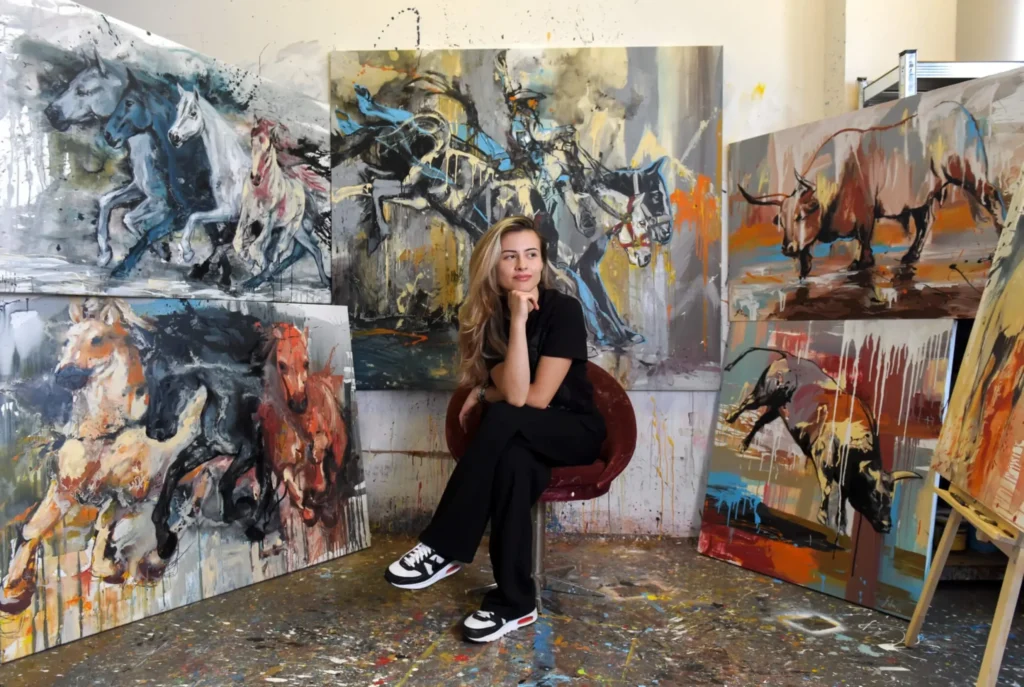 SILVI The Artist posing thoughtfully in her vibrant art studio surrounded by large expressive paintings of horses and bulls