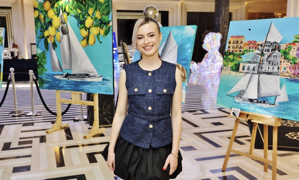 SILVI The Artist standing at her art exhibition surrounded by contemporary paintings featuring sailboats and Mediterranean-inspired scenes.